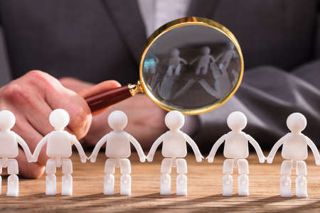 Close-up Of A Businessperson's Hand Holding Magnifying Glass Over Red Human Figure On Wooden Deskの写真素材