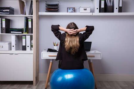 Rear View Of A Businesswoman Sitting On Fitness Ball Stretching Her Armsの写真素材