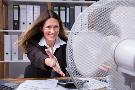 Happy Mature Businesswoman Cooling Herself With Electrical Fan At Workplaceの写真素材