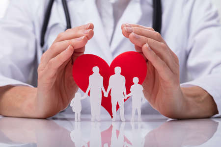 Close-up Of A Doctor's Hand Protecting Family Cut Out With Red Heart Shapeの写真素材