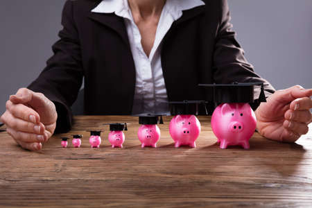 Human Hand Protecting Rising Pink Piggybanks With Graduation Hat Over Wooden Deskの写真素材