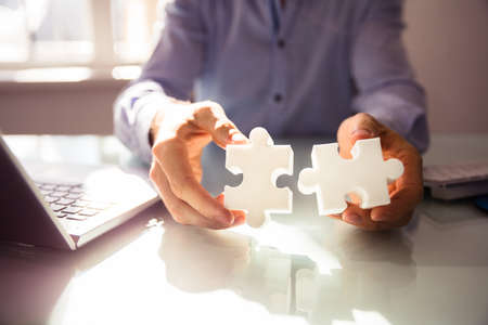 Close-up Of Businessperson's Hand Holding Two White Jigsaw Puzzle Over Deskの写真素材
