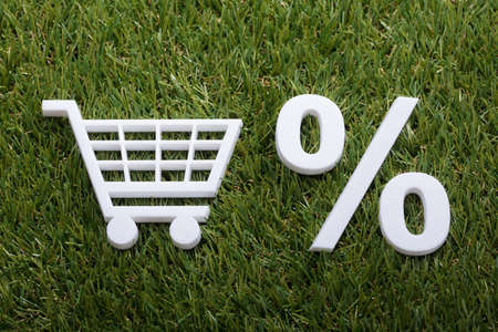 High Angle View Of White Miniature Shopping Cart And Percentage Sign On Green Grass