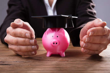 Close-up Of A Businessperson's Hand Protecting Pink Piggybank With Graduation Hat