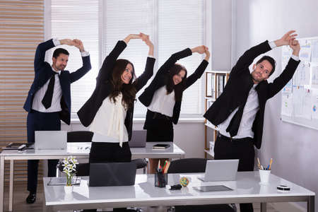 Happy Businesspeople Doing Stretching Exercise Behind Desk At Workplace