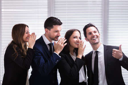 Businesspeople Whispering Into Young Male Colleague's Ear In Office