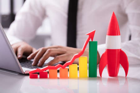 Rocket And Multi Colored Graph With Arrow In Front Of Businessperson Working On Laptop
