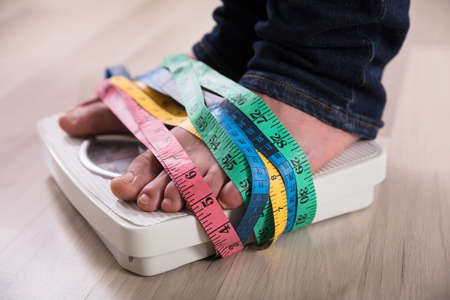 Close-up Of A Person's Feet On Weight Scale Wrapped With Multi Colored Measuring Tapeの写真素材