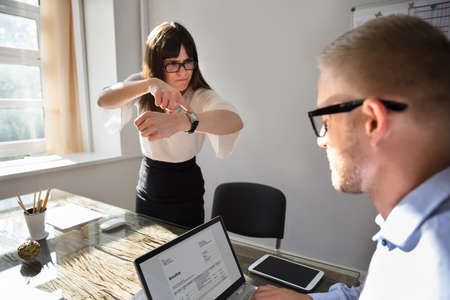 Angry Young Businesswoman Pointing On Wrist Watch While Colleague Checking Invoice On Laptop
