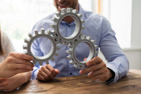 Close-up Of Two Businesspeople Connecting Gears Over Wooden Desk In Officeの写真素材