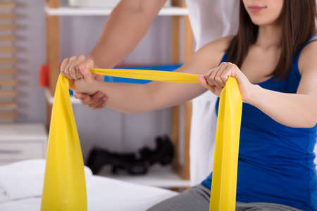 Physiotherapist Assisting Woman While Doing Exercise With Yellow Exercise Bandの写真素材