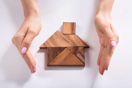 Close-up Of A Woman's Hand Protecting House Made Of Wooden Tangram Puzzle Over Deskの写真素材