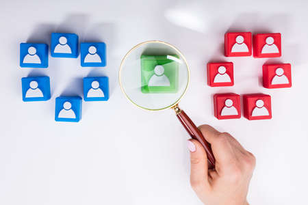 Businesswoman's Hand Holding Magnifying Glass Over Green Block Besides Team Of Red And Green Blocks