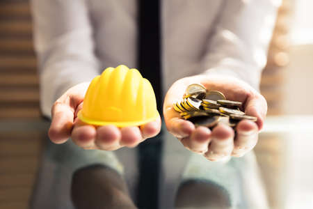 Human Hand Holding Yellow Hardhat And Many Golden Coins Over Deskの写真素材