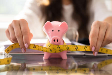 Businesswoman's Hand Measuring Piggybank With Yellow Measuring Tape On Glass Deskの写真素材