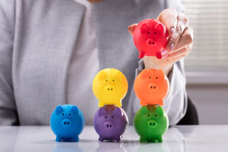 Close-up Of A Woman's Hand Stacking Multi Colored Piggybank On Deskの写真素材