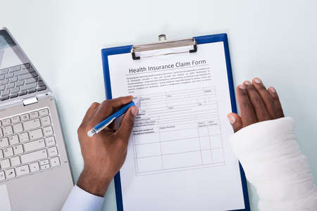 Overhead View Of Injured Man With Bandage Hand Filling Insurance Claim Form On Clipboardの写真素材
