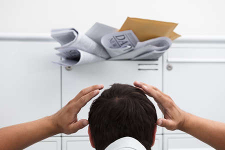 Close-up of man with hand on head in front of overloaded mailbox with junk mailの写真素材