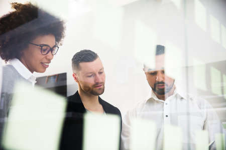 Group Of Young Businesspeople Sticking Adhesive Notes On Transparent Glass Wall In Officeの写真素材
