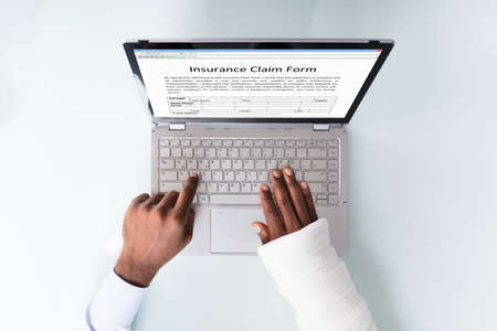 Overhead View Of A Businessman With Bandage Hand Filling Insurance Claim Form On Laptopの写真素材