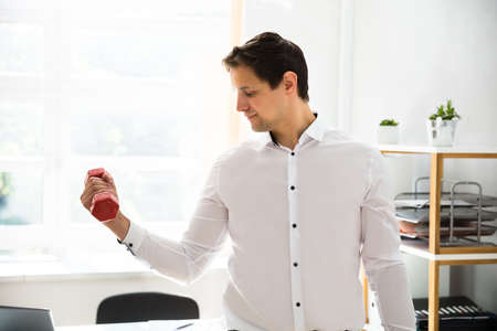 Young businessman exercising with red dumbbell in officeの写真素材