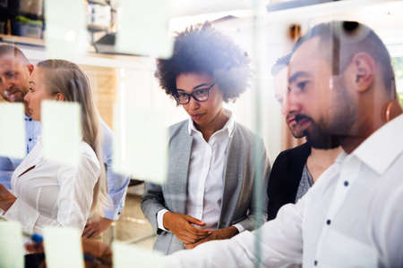 Group Of Young Businesspeople Sticking Adhesive Notes On Transparent Glass Wall In Office