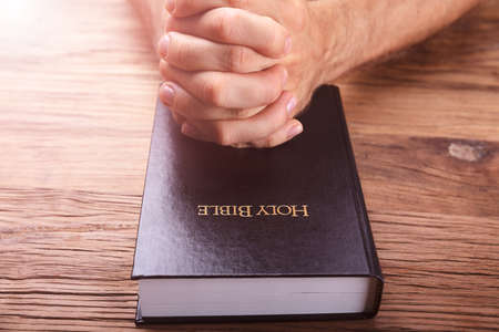 Man's Praying Hands On Bible Over Wooden Background