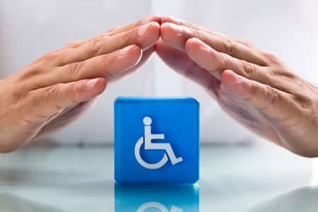 Close-up of a human hand protecting blue cubic block with handicap icon