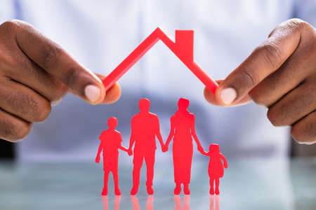 Close-up Of Businessman's Hand Holding Roof Over The Red Family Standing On Reflective Deskの写真素材