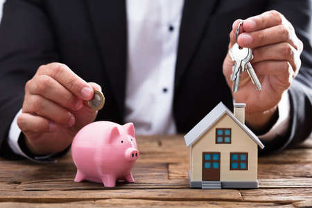 Businessperson's hand holding coin and key over piggybank and house on wooden deskの写真素材