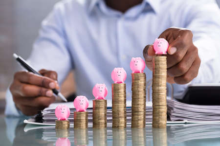 Close-up Of A Person's Hand Placing Pink Piggybank On Increasing Order Of Stacked Coinsの写真素材