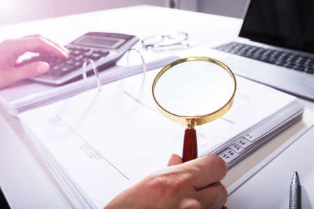 Close-up Of A Businessperson's Hand Analyzing Bill With Magnifying Glass In Officeの写真素材