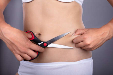 Midsection View Of A Woman's Hand Cutting Belly Fat With Scissorsの写真素材