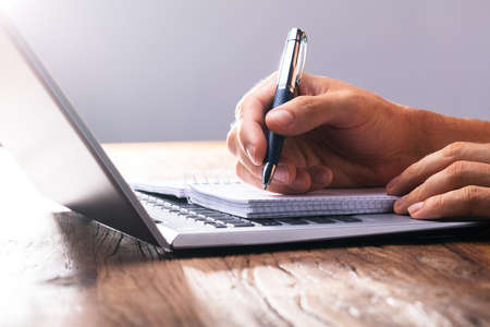 Close-up Of A Person's Hand Writing On Spiral Book Over Laptop
