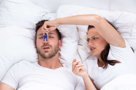 Frustrated Young Woman Trying To Stop Man's Snoring With Clothespin On Bed
