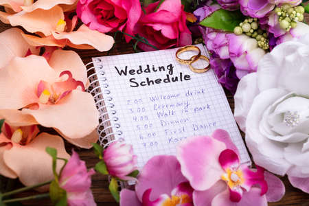 Two Golden Rings With Wedding Schedule On Spiral Notepad Surrounded By Flowers