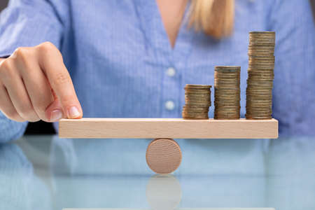 Businesswoman Balancing Stacked Coins With Finger On Wooden Seesaw