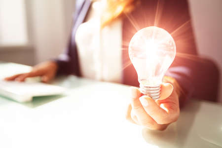 Close-up Of A Businesswoman's Hand Holding Illuminated Light Bulb In Office