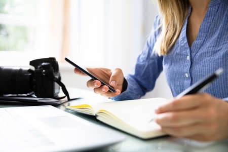 Female Editor's Hand With Mobile Phone Writing Schedule In Diary