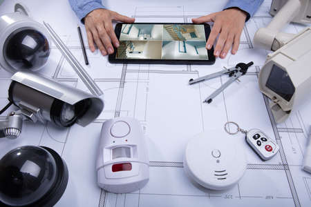 Architecture Looking At CCTV Footage On Digital Tablet With Various Security Equipment On Desk
