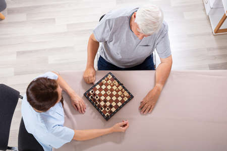 High Angle View Of Female Caretaker Playing Chess With Senior Man On Desk