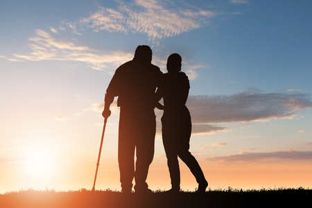 Silhouette Of Woman Assisting Her Disabled Father Walking In Park At Duskの写真素材