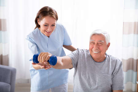 Young Female Nurse Helping Happy Senior Man With Dumbbell Exerciseの写真素材