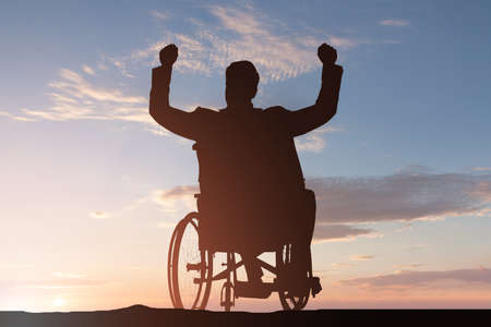 Silhouette Of A Disabled Man On Wheelchair With Arm Raised At Sunsetの写真素材
