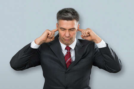 Portrait Of A Mature Businessman Covering His Ears With Fingers On Gray Backgroundの写真素材