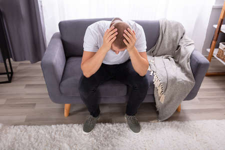 Young Man Sitting On Sofa Suffering From Headache
