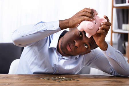 African Man Checking Savings In Piggybank With Coins On Wooden Deskの写真素材
