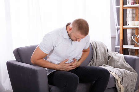 Young Man Sitting On Sofa Having Stomach Pain At Home