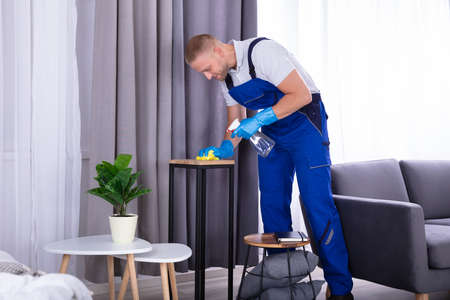 Young Male Janitor Wiping Wooden Furniture In House
