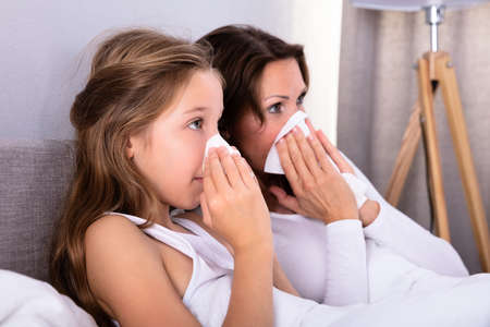 Mother And Daughter Suffering From Cold Blowing Her Nose With Handkerchief On Bed